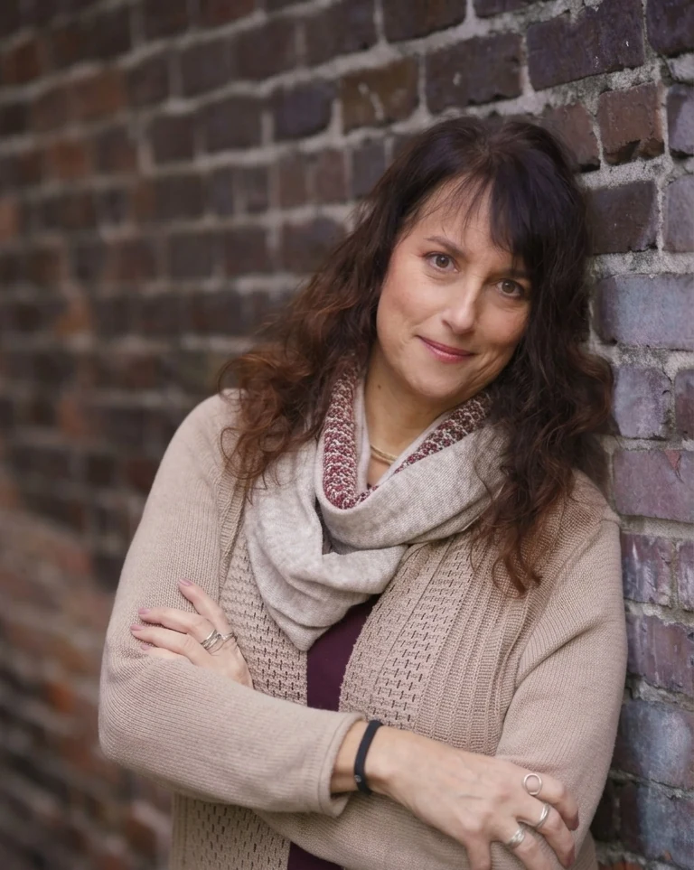 Issy Kleiman, MA, LMFT, AAMFT Approved Supervisor, standing with her arms crossed against a brick wall in a camel cardigan